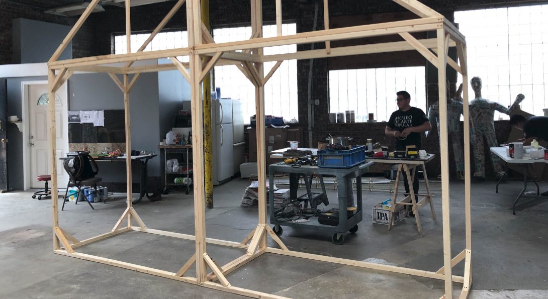 A photograph of a wooden structure inside a large construction room. The wooden structure has the outline of a house with a triangle-shaped roof.