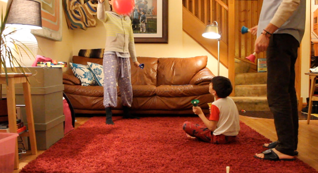 A photograph of a video still from Sensitive Equipment. The video still is in a living room setting. A young person is sitting on a red carpet on the floor. They are looking upwards at another young person