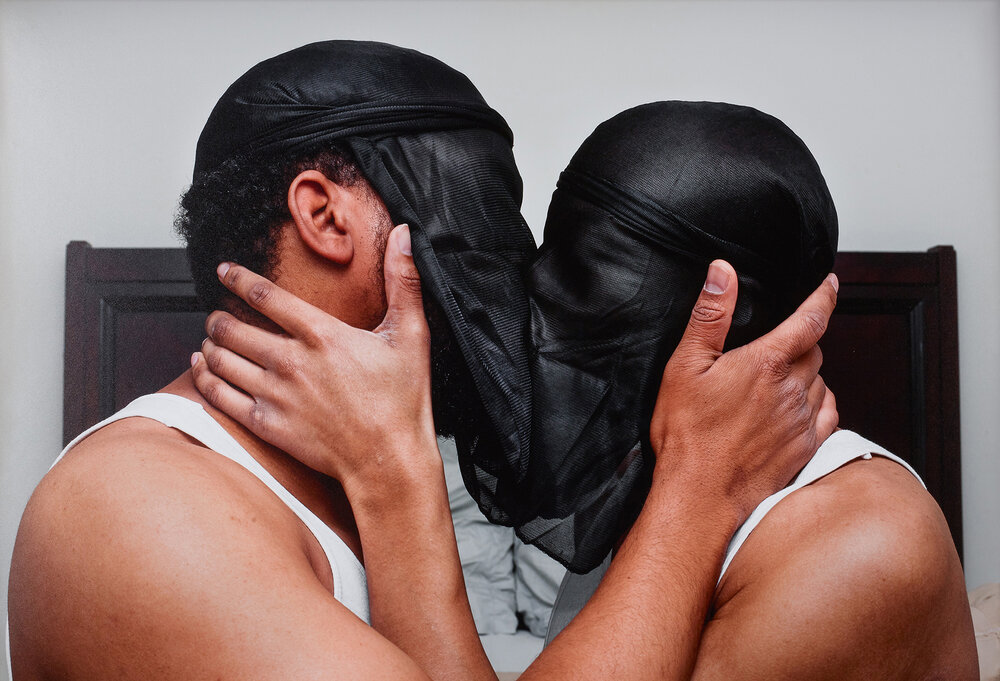 Two black men wearing black doo-rags around the front of their faces kiss while holding each other's faces.|A black man wearing only strips of red and white fabric around his arms and legs stands on a wooden box in front of white screen. He is posed on one leg