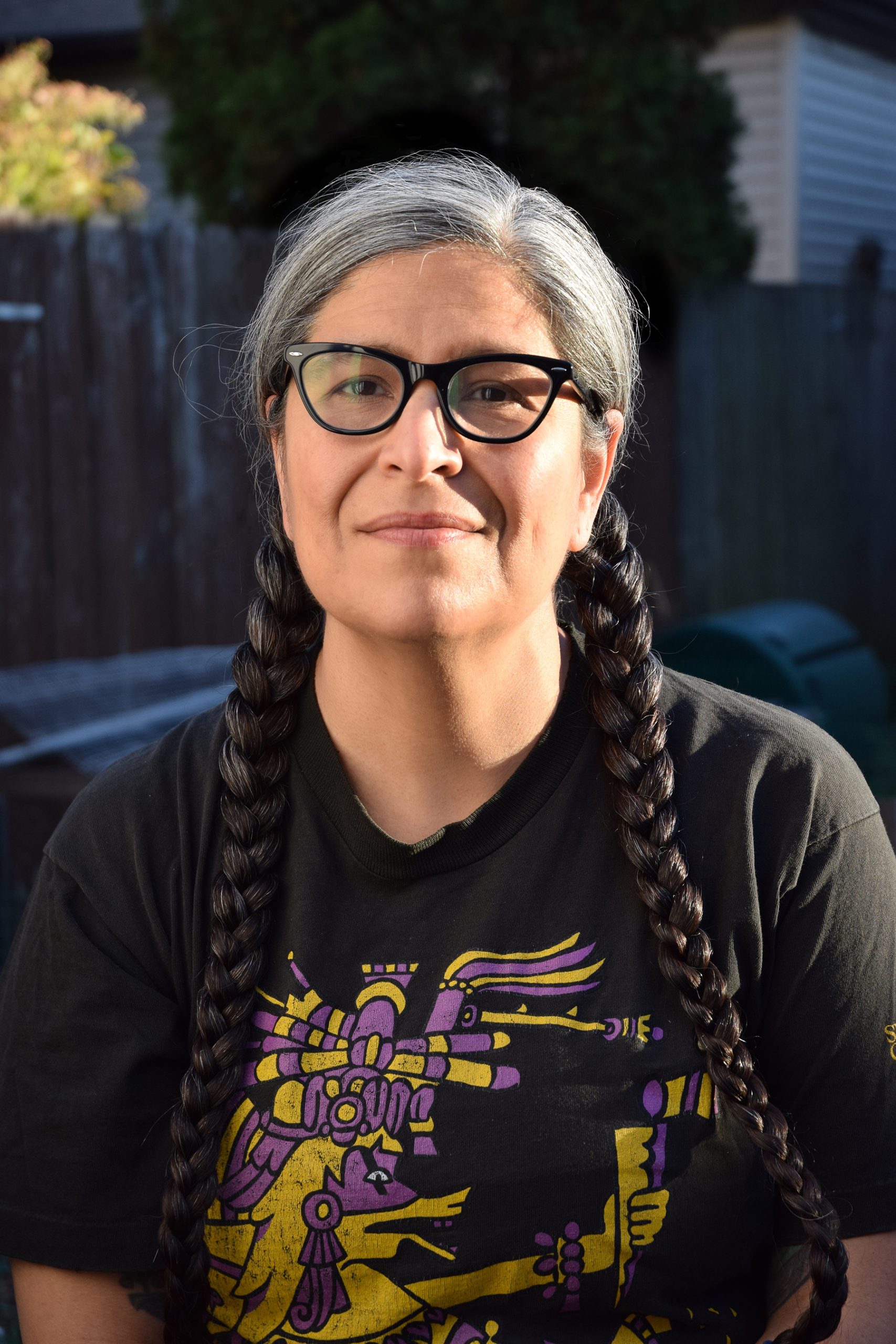 Headshot of a medium-skinned woman with glasses and long grew and black hair in two braids.|Two people sitting with their backs to the camera. They are in conversation and there is a tv with black and white footage on it. The rest of the photograph is orange tinted.|Four black and white photographs are spread out on a hard surface.