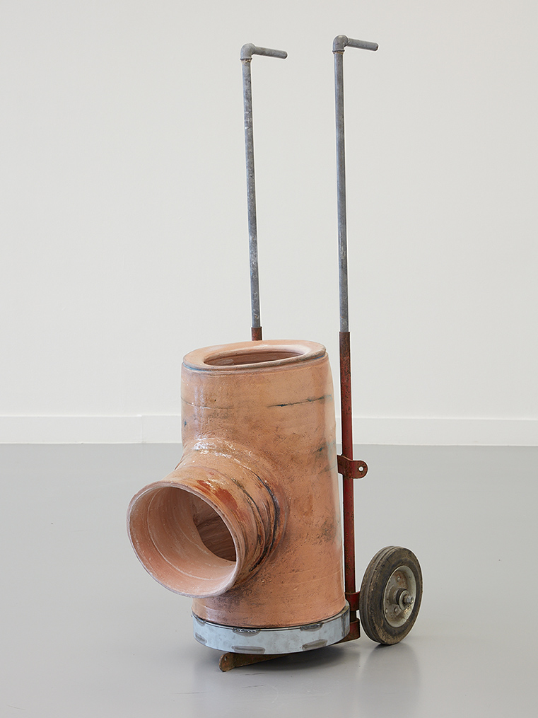 A sculpture in a white walled space. The object is made of a tube with a circular opening in the front and two pipes stretched up the back. The object is on wheels.|A medium skinned person with brown curly hair and wearing a pink top. They lean on a table and smile